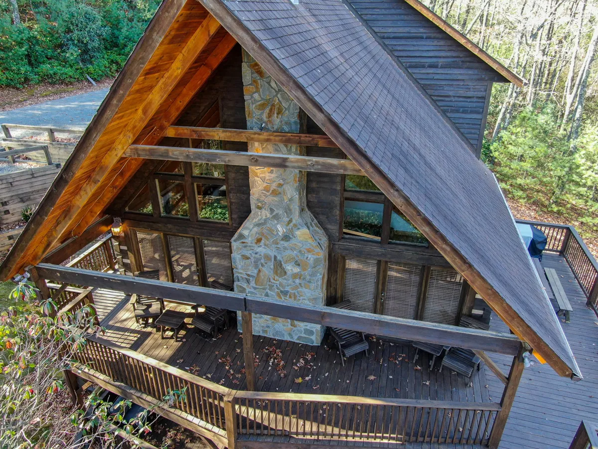 This image showcases the wooden deck and stone chimney of Happy Trees Cabin, surrounded by lush trees. The spacious deck offers room for gathering, relaxing, and enjoying meals outdoors, while the large windows invite natural light and connect the interior to the beautiful surroundings.