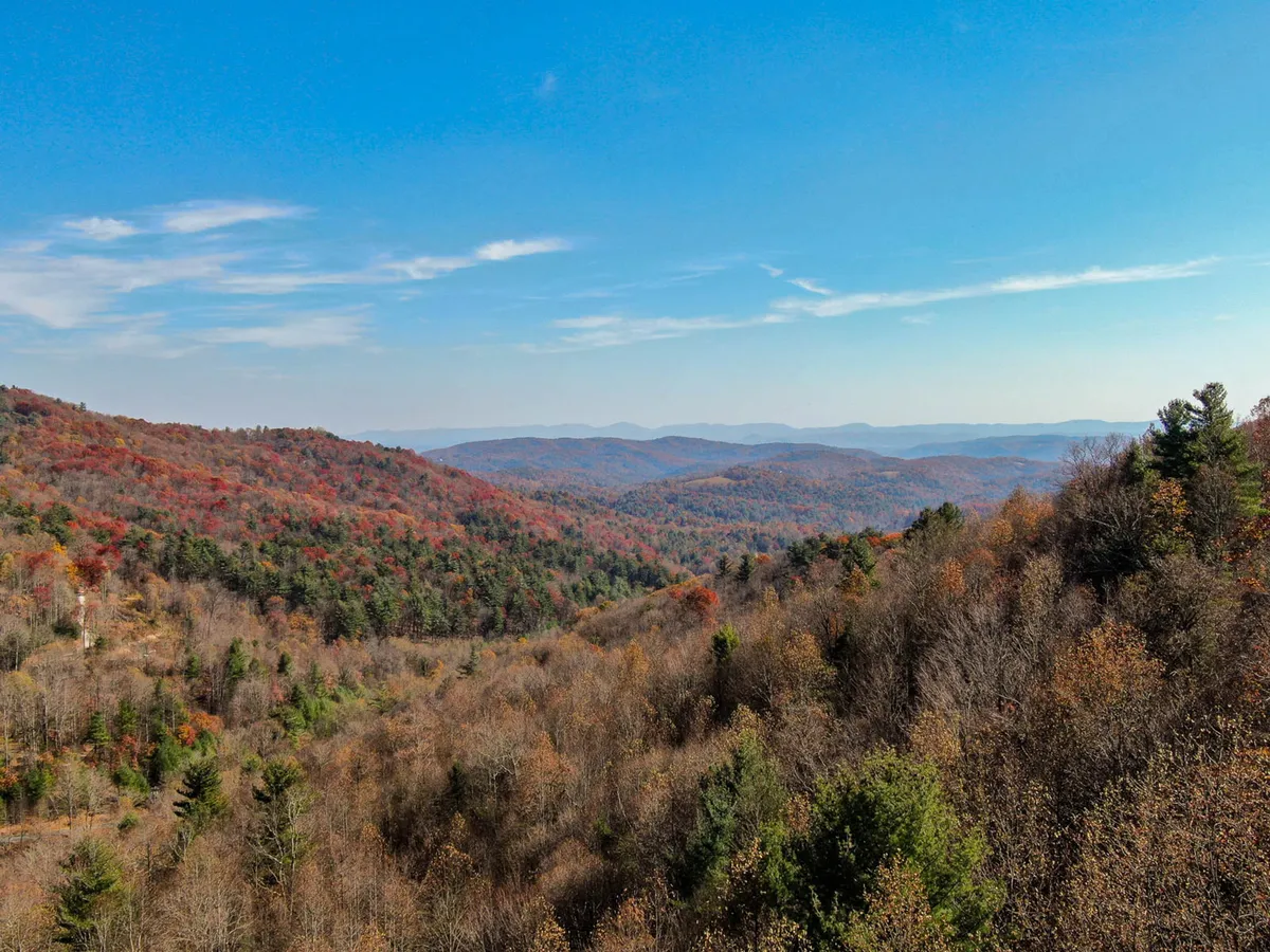 A panoramic view of rolling hills covered in autumn foliage, with distant mountains rising beyond. The tranquil landscape bathes in warm golden light, ideal for sunrise hikes, wildlife observation, and appreciating the seasonal beauty of the Blue Ridge Parkway.