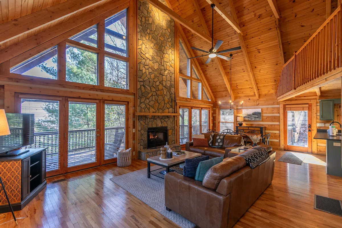 The image showcases the inviting great room of Happy Trees Cabin, featuring a stunning floor-to-ceiling stone fireplace, soaring exposed wooden beams, and expansive windows that flood the space with natural light. Plush sofas create a cozy, welcoming seating area ideal for gathering with family or friends.