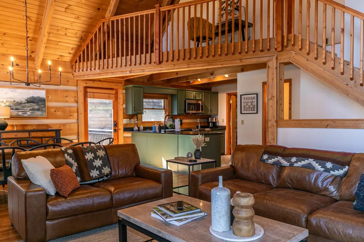 A wide-angle perspective capturing the spacious interior with leather couches, a wooden dining table for eight, an open-plan kitchen, and lofted sleeping area above. The arrangement emphasizes how the living, dining, and kitchen spaces seamlessly connect, with warm wooden finishes throughout.
