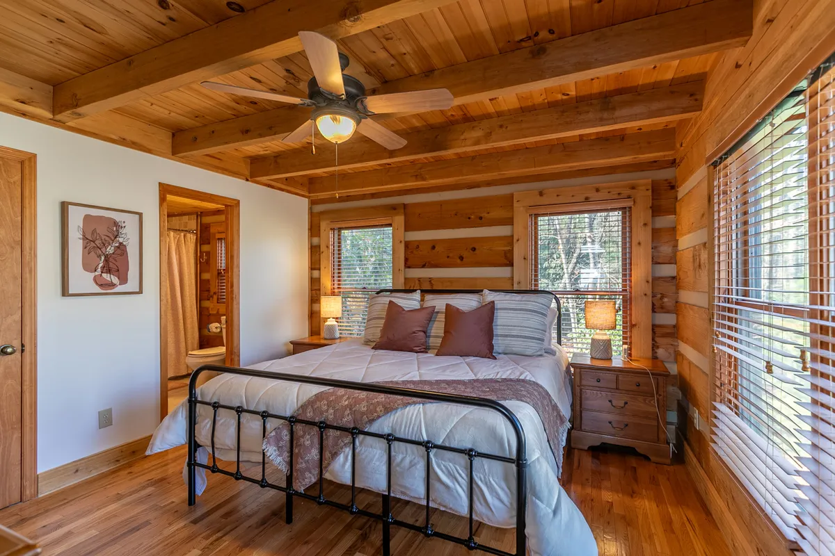 A serene bedroom featuring a comfortable bed beneath a wooden ceiling, with softly diffused natural light streaming through windows. The space is enhanced by a nightstand and lamp, evoking a peaceful, restful environment ideal for relaxation and unwinding after a day outdoors.