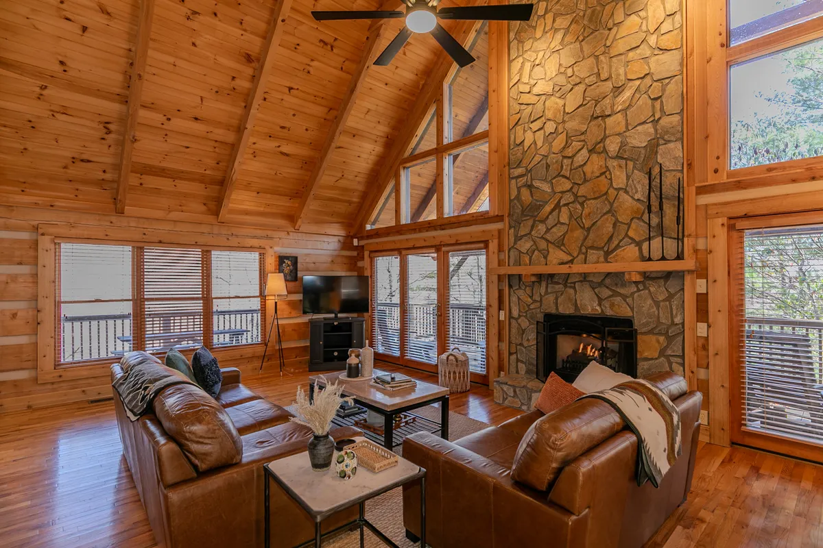 This image features the heart of the great room: a grand stone gas fireplace framed by soaring wooden beams and a wall of windows that let in abundant natural light. Plush leather sofas and a cozy coffee table invite relaxation and conversation, while the view out the windows provides a strong sense of connection to the surrounding forest.