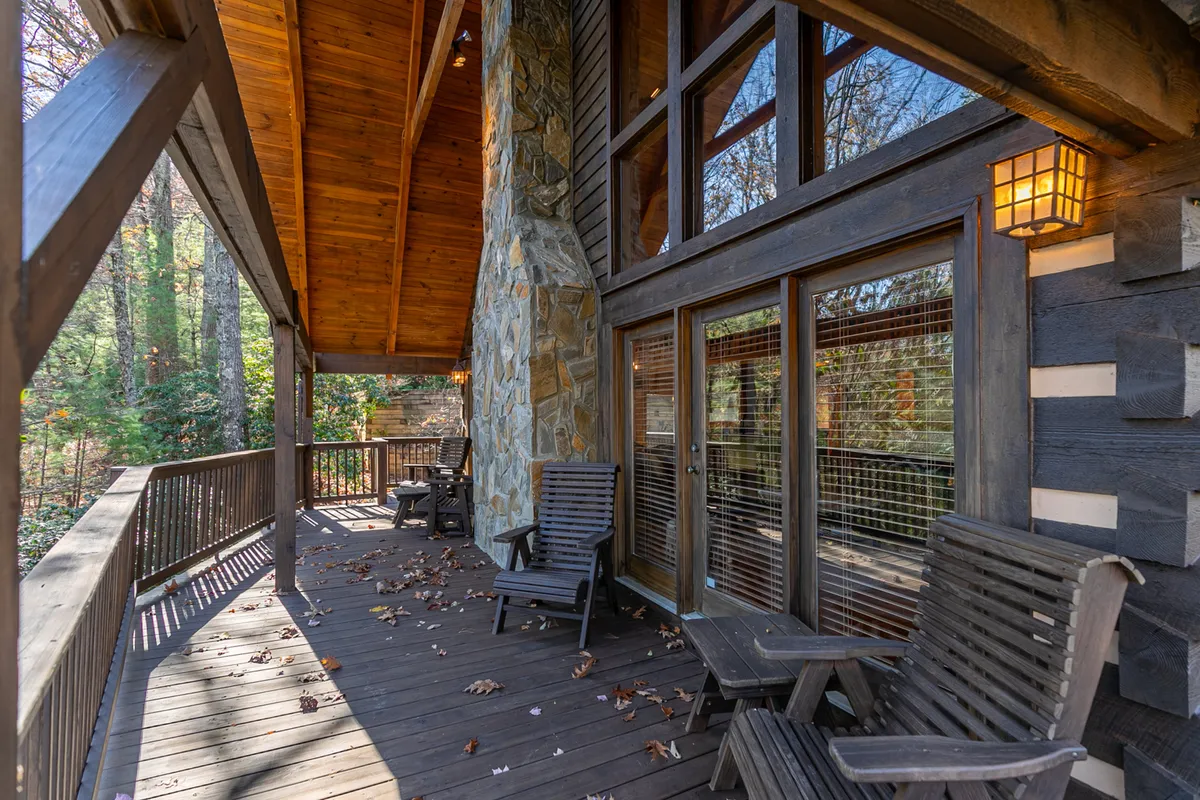 The deck, bordered by a stone wall and large windows, is furnished with comfortable wooden chairs, offering a cozy spot for reading or socializing while surrounded by tranquil forest views. The inviting ambiance is ideal for peaceful mornings or stargazing nights.