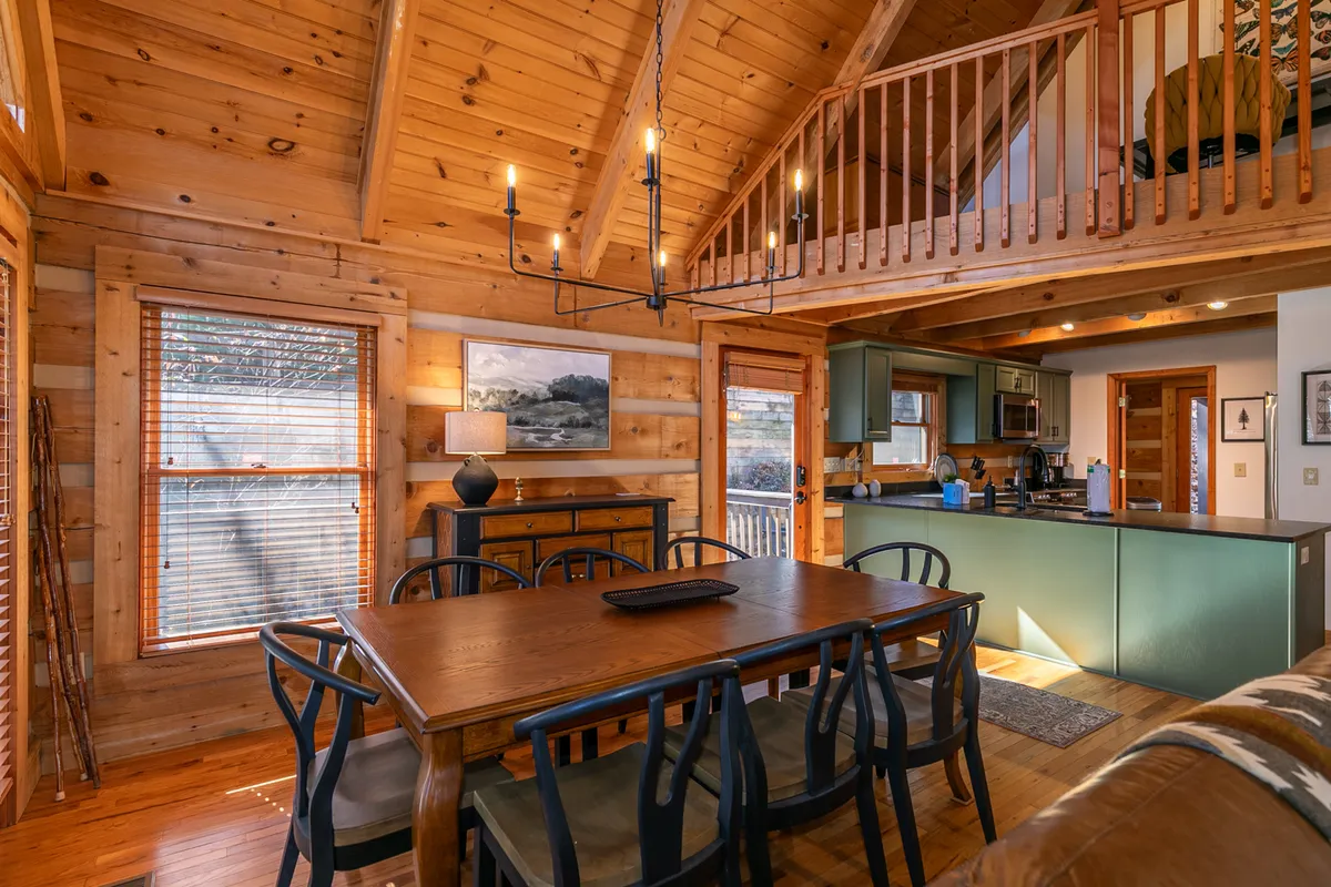 A bright dining area with a large wooden table, stylish chandelier, open kitchen, and large windows that frame forest views. The space feels inviting, filled with natural light, and visually connects to the adjacent great room.