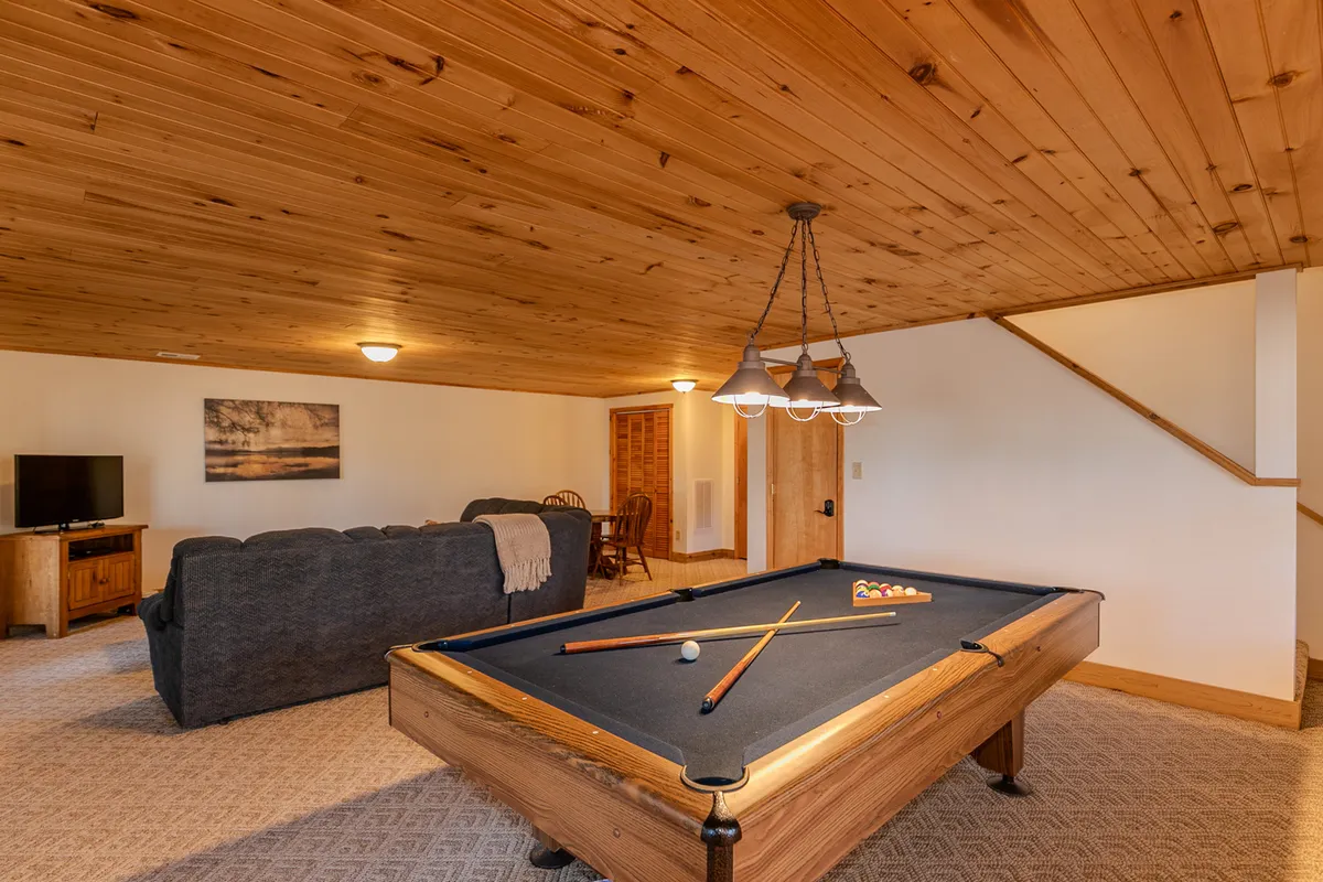 A vibrant game room featuring a pool table as the focal point, flanked by a comfortable sofa and a large television. Overhead lighting sets a casual, fun atmosphere perfect for both friendly competition and movie nights. The room is clearly designed for group entertainment and relaxation.