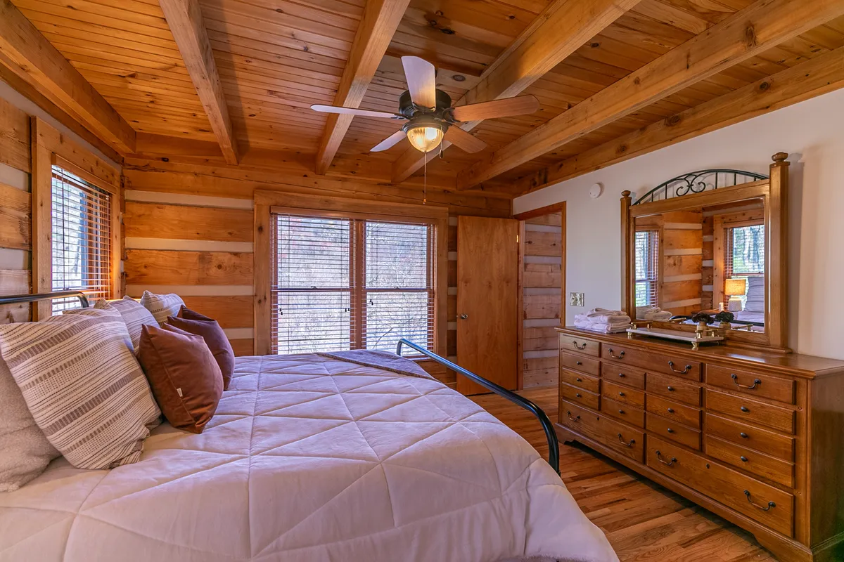 An inviting upstairs bedroom with a bed adorned in decorative pillows, a wooden dresser, ceiling fan, and large windows that fill the space with natural light. The overall vibe is warm and welcoming, perfect for guests who enjoy mountain air and morning breezes.