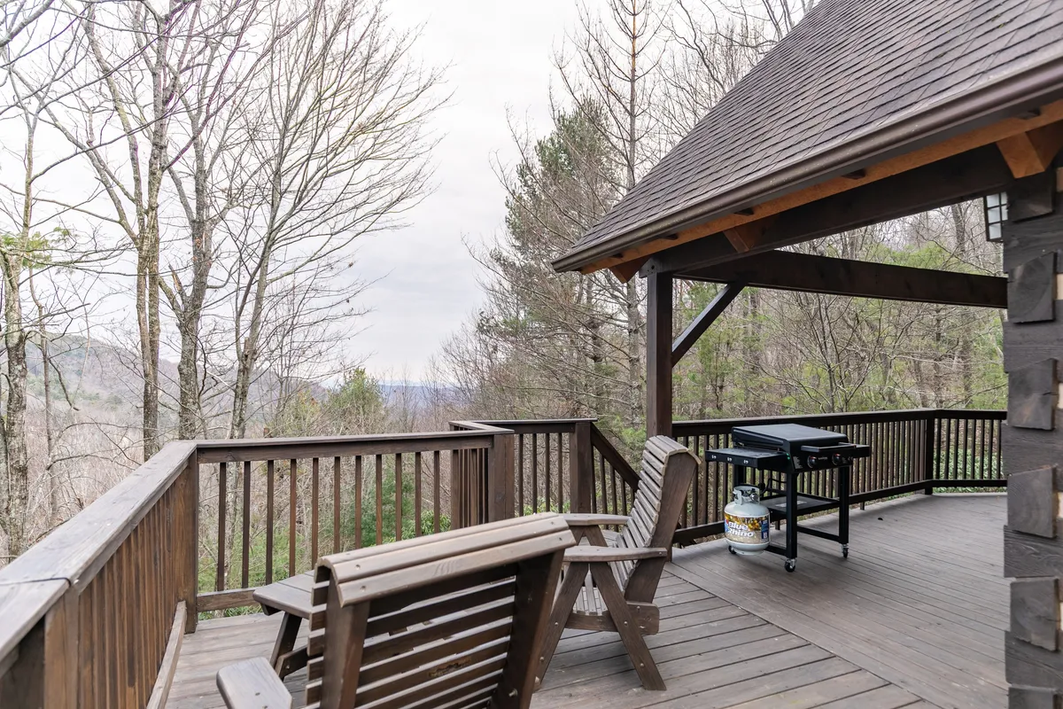 A wide wooden deck stretches out with a gas grill on one side, outdoor seating, and an expansive view of the mountains visible beyond bare trees. The space invites relaxation and alfresco dining with nature as the backdrop.